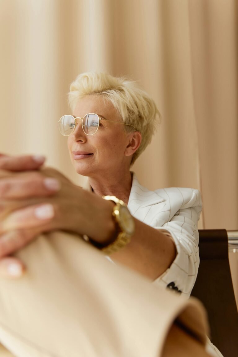 Elegant middle-aged woman in a business suit and glasses sitting confidently indoors.